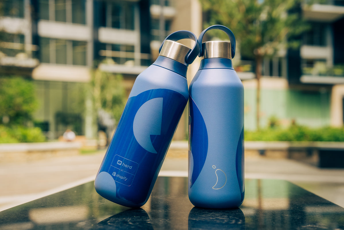 Two blue Herd Chilly's water bottles with Herd logo on a reflective surface in an outdoor setting.